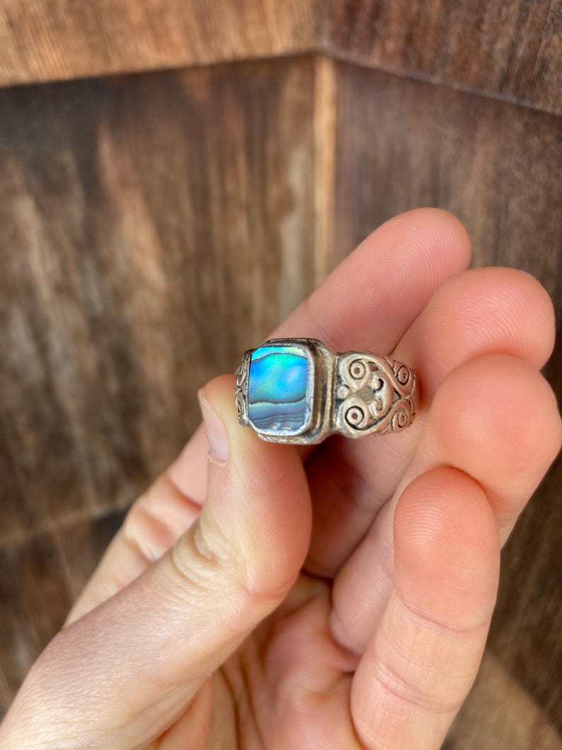 May include: A silver ring featuring a square abalone shell inlay. The ring band has a decorative, swirling design.