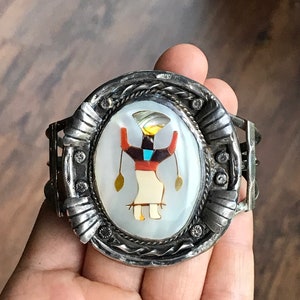 May include: Silver cuff bracelet with an oval centerpiece featuring a Native American figure inlaid with mother-of-pearl, coral, and turquoise. The bracelet has a detailed, textured design and is held in a hand.