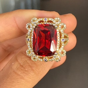 May include: A gold ring with a large, red, rectangular gemstone surrounded by smaller, clear gemstones. The ring has a detailed, ornate design.