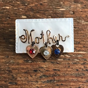 May include: A white mother's day pin with the word "Mother" in cursive script and three dangling hearts with red, clear, and blue rhinestones.