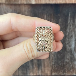 May include: A gold filigree ring with a rectangular design. The ring features a delicate, intricate pattern of swirls and curves.