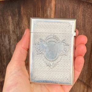 May include: A silver cigarette case with an intricate engraved design. The case has a rectangular shape and a hinged lid. The engraving features a central oval surrounded by a floral motif.