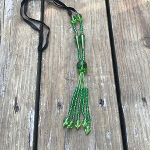 Op de afbeelding: Een groene kralenketting met een zwart lint. De ketting heeft een kwast van groene kralen aan de onderkant.
