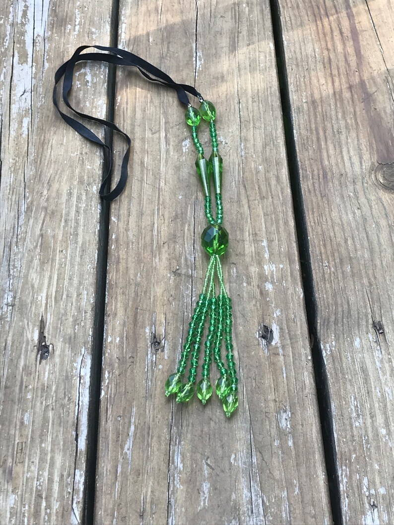 Op de afbeelding: Een groene kralenketting met een zwart lint. De ketting heeft een kwast van groene kralen aan de onderkant.