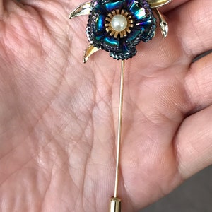Peut inclure: Une broche en forme de fleur dor&eacute;e avec des p&eacute;tales bleu et vert iris&eacute;s et un centre en perle. La broche est fix&eacute;e &agrave; une longue &eacute;pingle dor&eacute;e.