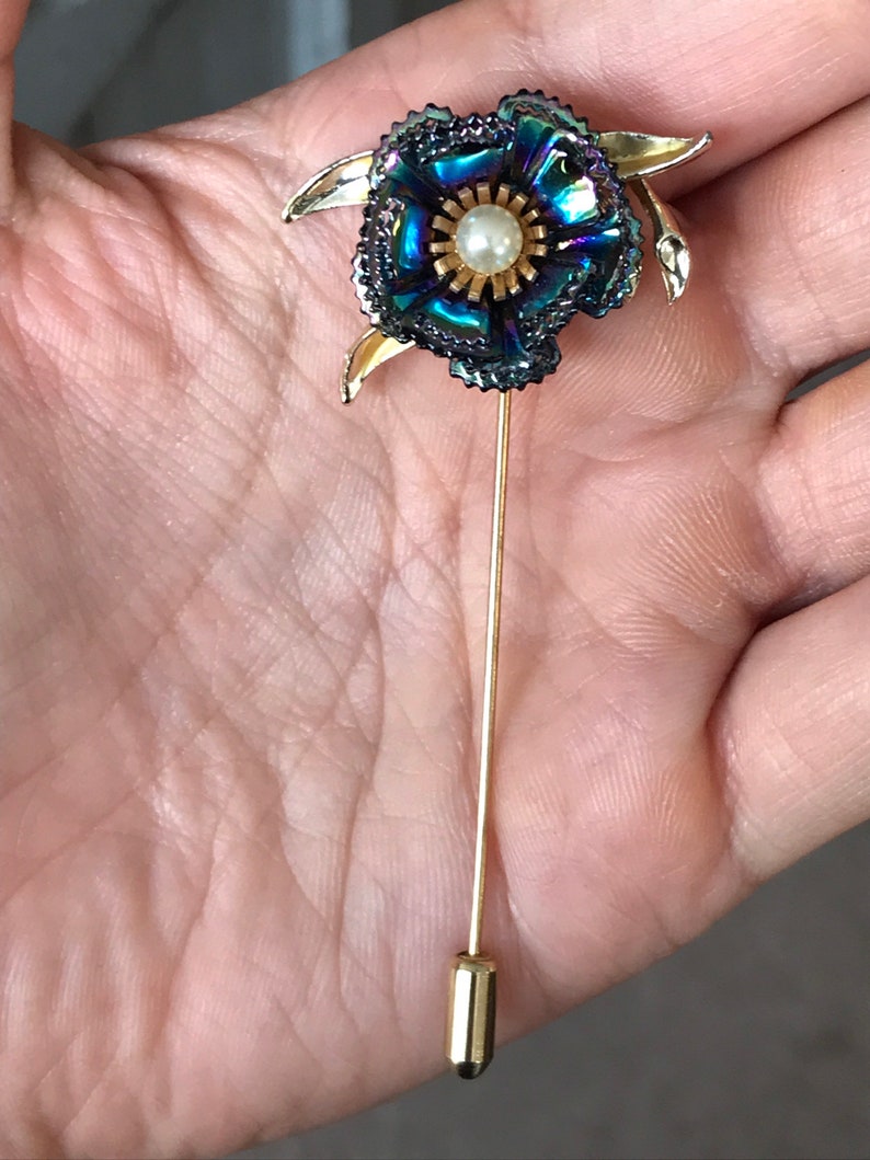 Peut inclure: Une broche en forme de fleur dor&eacute;e avec des p&eacute;tales bleu et vert iris&eacute;s et un centre en perle. La broche est fix&eacute;e &agrave; une longue &eacute;pingle dor&eacute;e.