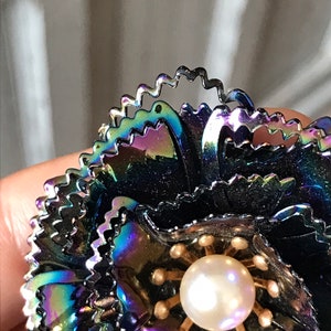 Peut inclure: Une broche en forme de fleur noire iris&eacute;e avec un centre en perle. La fleur est compos&eacute;e de plusieurs couches de p&eacute;tales m&eacute;talliques &agrave; bord festonn&eacute;.