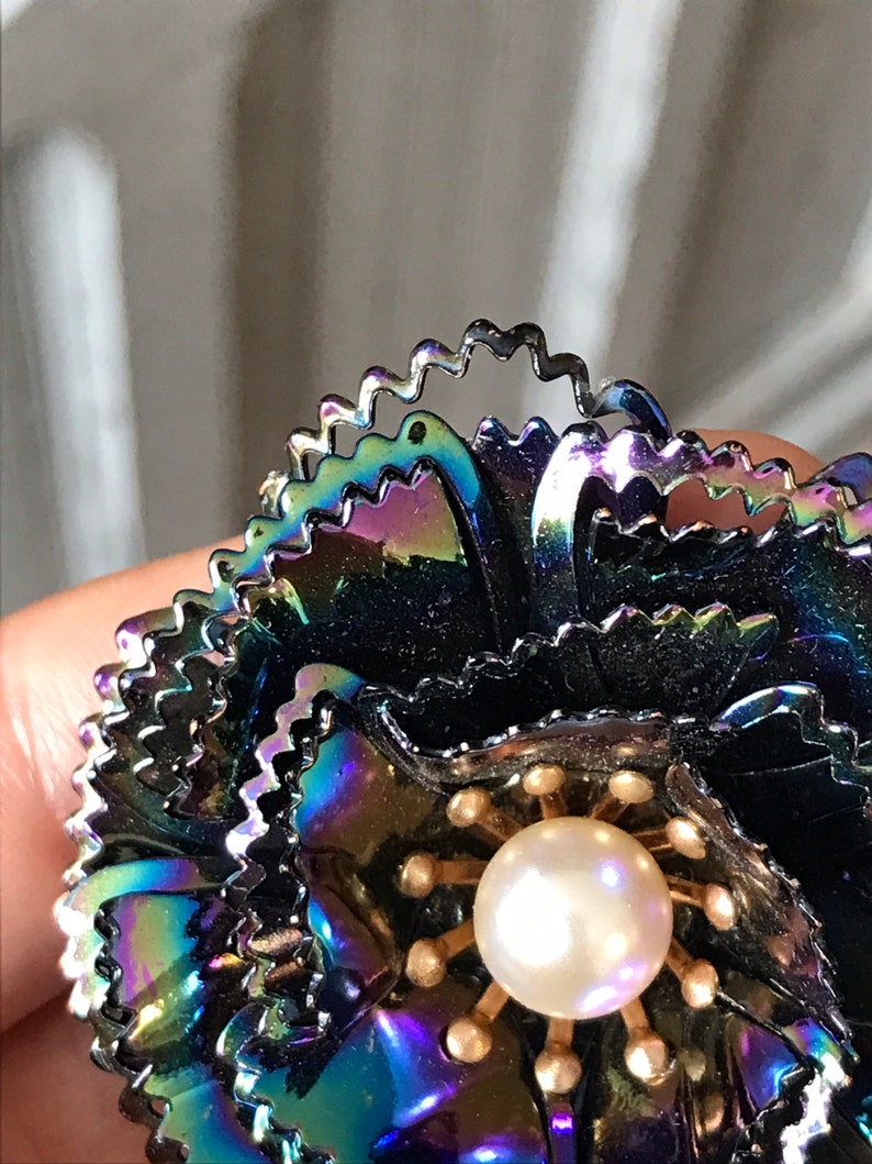 Peut inclure: Une broche en forme de fleur noire iris&eacute;e avec un centre en perle. La fleur est compos&eacute;e de plusieurs couches de p&eacute;tales m&eacute;talliques &agrave; bord festonn&eacute;.