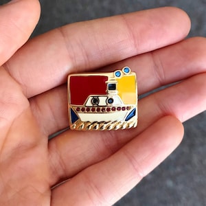 May include: A gold-toned enamel pin featuring a stylized boat with red, yellow, blue, and white accents. The boat has two small blue rhinestones on the top.