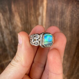 May include: A silver ring with an abalone shell inlay. The ring has a detailed design with swirls and a rectangular shaped stone.