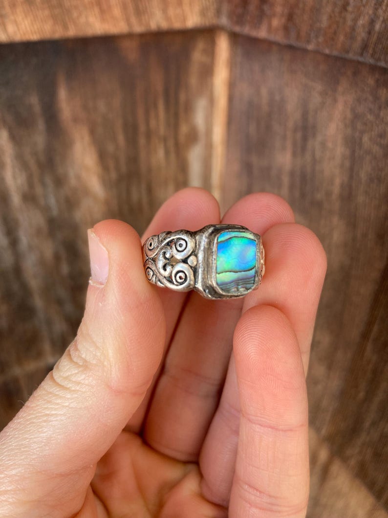 May include: A silver ring with an abalone shell inlay. The ring has a detailed design with swirls and a rectangular shaped stone.