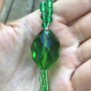 Op de afbeelding: Een groene glazen kralenketting met een grote, gefacetteerde, groene glazen hanger. De ketting is gemaakt van kleine, ronde kralen en heeft een kwast aan de onderkant.