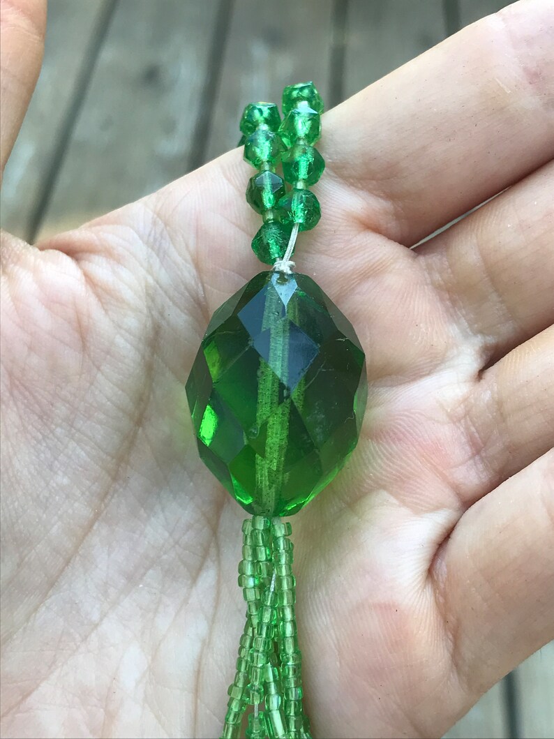 Op de afbeelding: Een groene glazen kralenketting met een grote, gefacetteerde, groene glazen hanger. De ketting is gemaakt van kleine, ronde kralen en heeft een kwast aan de onderkant.