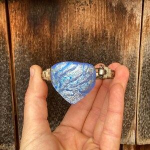 May include: A dichroic glass bracelet with a blue and silver heart-shaped centerpiece. The glass has a textured, shimmering appearance. The bracelet's metal band is silver-toned and woven. The background is a wooden surface.