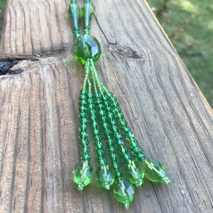 Op de afbeelding: Een groene kralenketting met een kwast. De ketting is gemaakt van kleine, gefacetteerde kralen en heeft een grote, gefacetteerde kraal aan de onderkant. De kwast is gemaakt van meerdere strengen kralen.