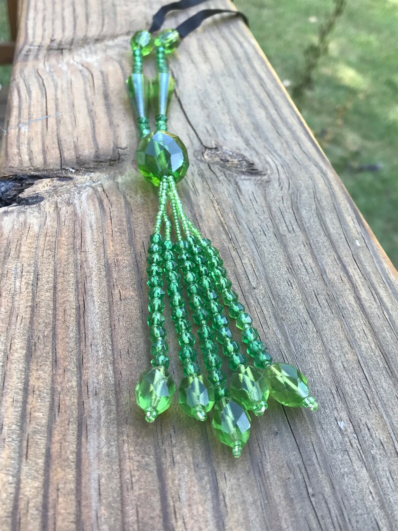 Op de afbeelding: Een groene kralenketting met een kwast. De ketting is gemaakt van kleine, gefacetteerde kralen en heeft een grote, gefacetteerde kraal aan de onderkant. De kwast is gemaakt van meerdere strengen kralen.