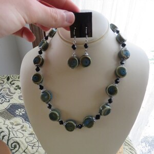 May include: A necklace and earring set displayed on a mannequin. The necklace and earrings feature olive green and blue ceramic beads, interspersed with black crystal beads. The earrings are dangling. The jewelry is displayed on a cream-colored bust.