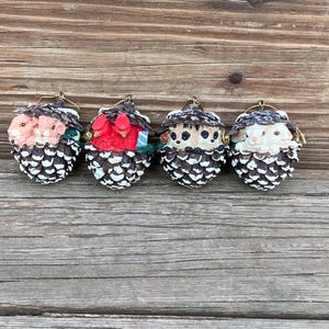 May include: Four small pine cone ornaments with different animal faces peeking out. The ornaments are white with brown accents and have a gold loop for hanging.