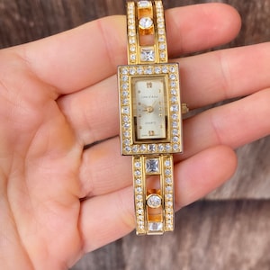 May include: Gold-tone wristwatch with a rectangular face and a crystal-encrusted band. The watch face has a white background with black Roman numeral hour markers and the words "Cole d'Azur" and "Quartz".
