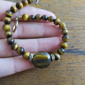 May include: A brown and gold tiger's eye beaded bracelet with a larger oval tiger's eye bead in the center. The bracelet has a gold clasp.