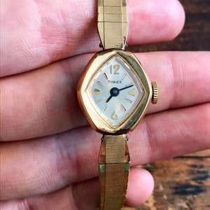 May include: A gold-toned Timex wristwatch with a white face and black hands. The watch has a diamond-shaped face and a gold-toned metal band.