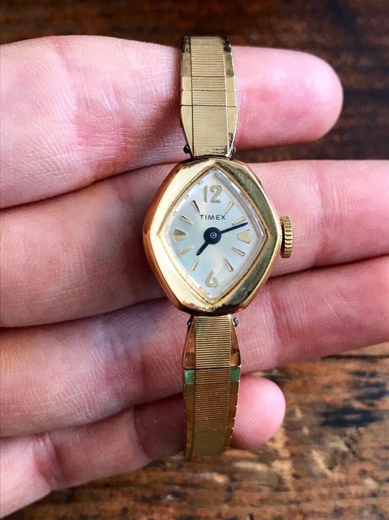 May include: A gold-toned Timex wristwatch with a white face and black hands. The watch has a diamond-shaped face and a gold-toned metal band.