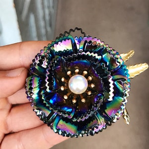 Peut inclure: Une grande broche en forme de fleur iris&eacute;e avec un centre en perle. La fleur est compos&eacute;e de plusieurs couches de p&eacute;tales m&eacute;talliques et a un centre dor&eacute;.