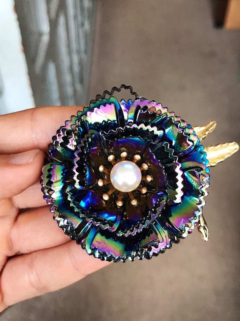 Peut inclure: Une grande broche en forme de fleur iris&eacute;e avec un centre en perle. La fleur est compos&eacute;e de plusieurs couches de p&eacute;tales m&eacute;talliques et a un centre dor&eacute;.