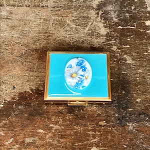 May include: A vintage compact with a blue enamel exterior and a gold trim. The compact features a floral design with white and blue flowers.