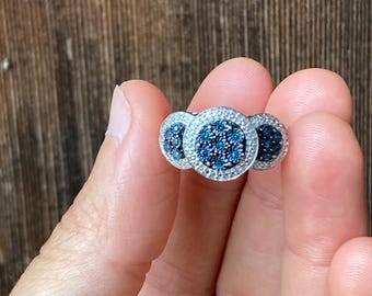 May include: A silver ring with three circular clusters. Each cluster features a dark blue center surrounded by small white stones. The ring is held up by a hand against a brown wooden background.