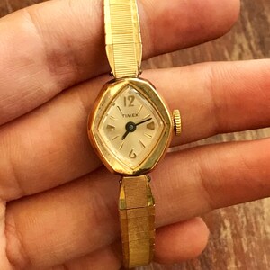 May include: A gold-toned Timex wristwatch with a white face and black hands. The watch has a diamond-shaped face and a gold-toned metal band.