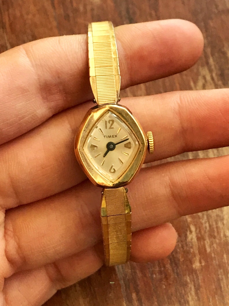 May include: A gold-toned Timex wristwatch with a white face and black hands. The watch has a diamond-shaped face and a gold-toned metal band.