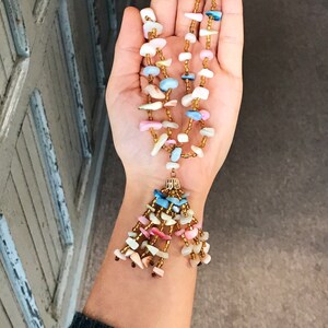 May include: A multi-strand necklace with a tassel made of small, colorful, and iridescent stones. The necklace is made of gold beads and has a variety of colors, including pink, blue, and white.