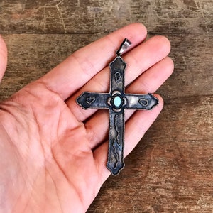 May include: A silver cross pendant with a turquoise stone in the center. The cross has a detailed design with a rope-like border.