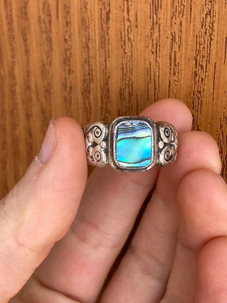 May include: A silver ring featuring a rectangular abalone shell inlay. The ring band has a decorative design.