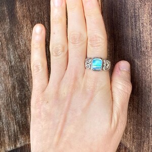 May include: A silver ring featuring a blue and green abalone shell inlay. The ring has a decorative, scroll-like design.
