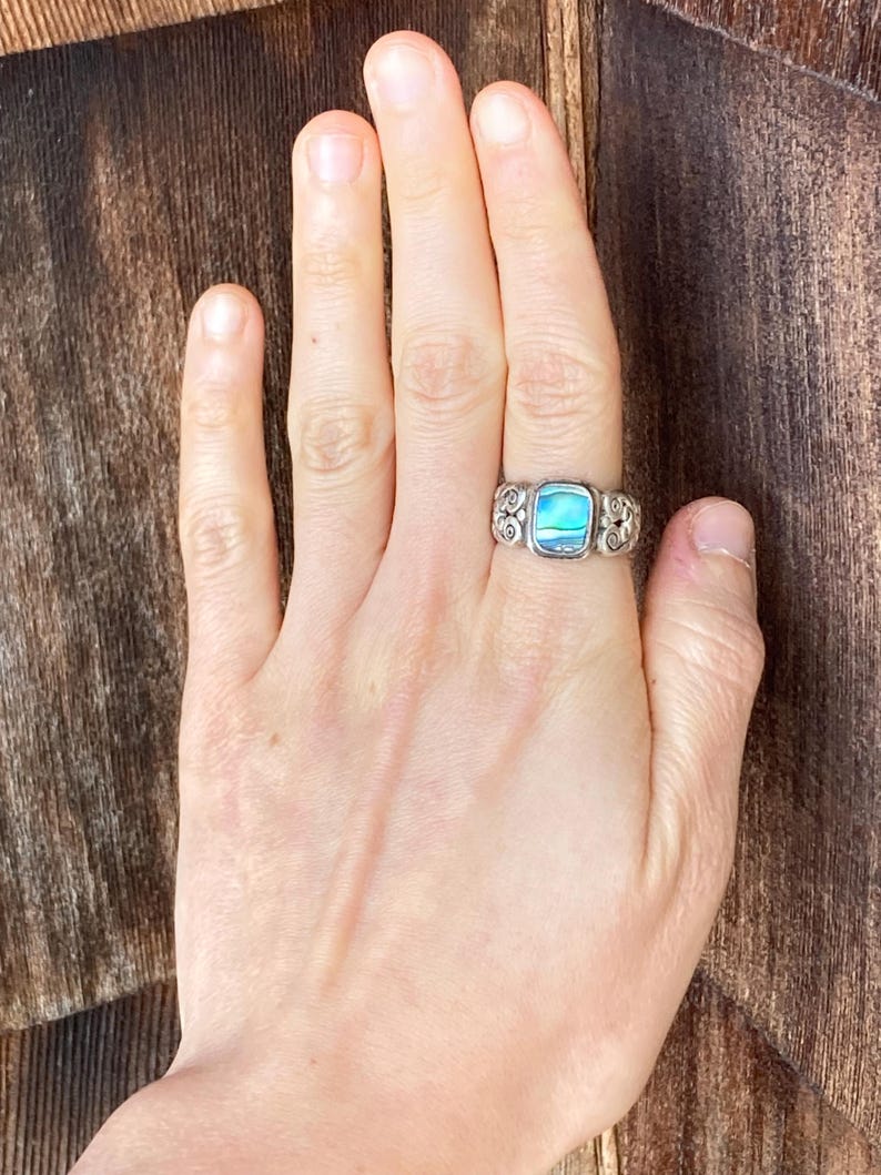 May include: A silver ring featuring a blue and green abalone shell inlay. The ring has a decorative, scroll-like design.