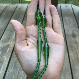 Op de afbeelding: Een lange groene glazen ketting met een grote traanvormige hanger. De ketting is gemaakt van gefacetteerde kralen en heeft een zwart koord.