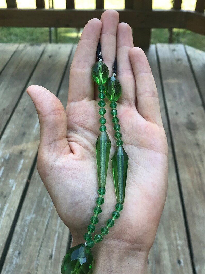 Op de afbeelding: Een lange groene glazen ketting met een grote traanvormige hanger. De ketting is gemaakt van gefacetteerde kralen en heeft een zwart koord.