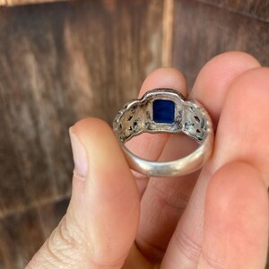 May include: A silver ring with a blue stone set in the centre. The ring has a detailed design around the stone.