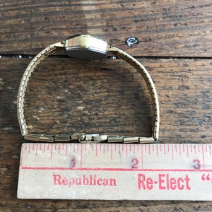 May include: A gold-toned wristwatch with a square face and a metal band. The watch is laid out on a wooden surface with a ruler next to it. The ruler has the words "Republican" and "Re-Elect" printed on it.