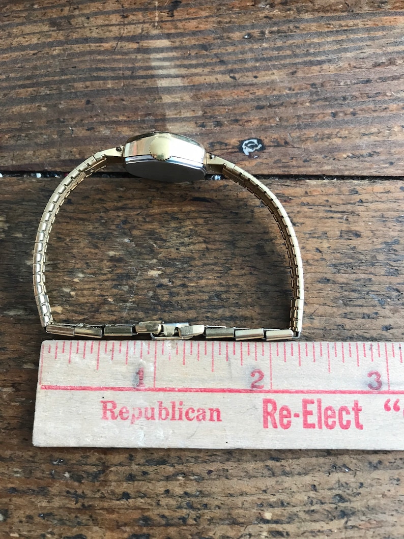 May include: A gold-toned wristwatch with a square face and a metal band. The watch is laid out on a wooden surface with a ruler next to it. The ruler has the words "Republican" and "Re-Elect" printed on it.
