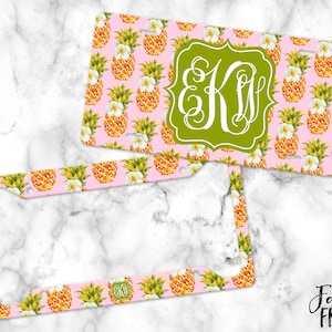 May include: A pink and white license plate frame with a pineapple pattern and a green monogram "EK" in the center. The frame is made of metal and has a shiny finish.