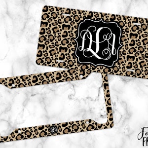 May include: A black and tan leopard print license plate frame with a monogram in a black and white circle. The monogram is the letters "QA".
