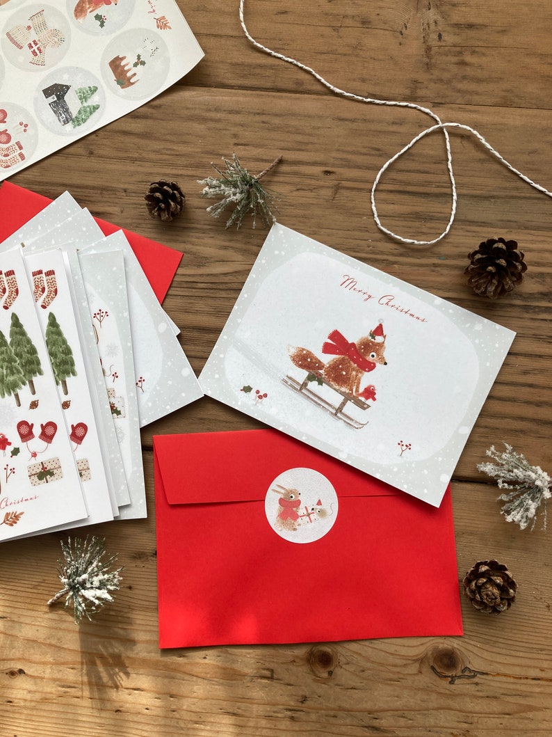 May include: A set of Christmas cards with a red envelope and a sticker featuring a fox and a rabbit on a sled. The card features a fox on a sled with the text "Merry Christmas".