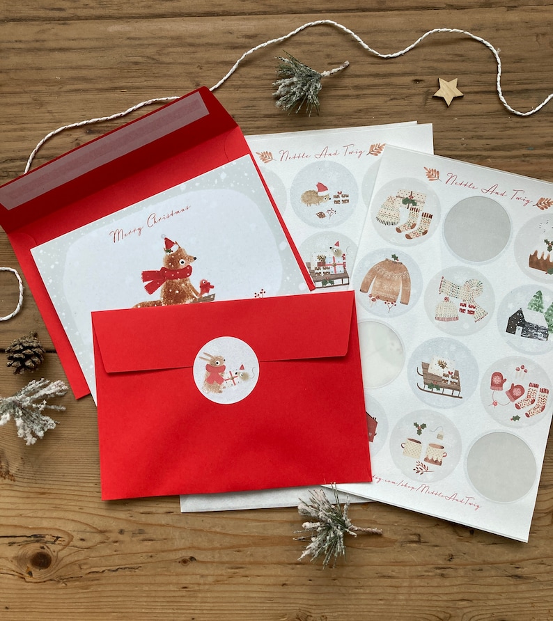 May include: A red envelope with a white card inside. The card has a drawing of a fox wearing a red hat and scarf. The text "Merry Christmas" is written on the card. The envelope has a sticker of a rabbit wearing a red hat and scarf. The image also shows a sheet of stickers with various winter-themed designs.