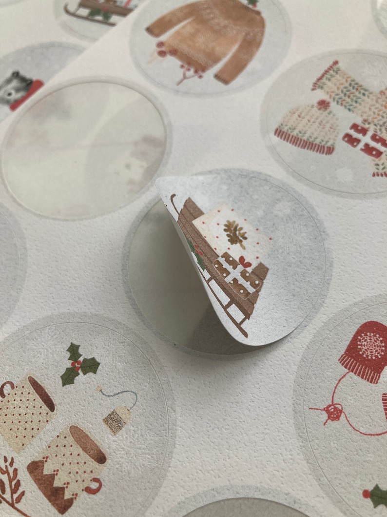 May include: A sheet of round, white stickers with various winter-themed illustrations. The stickers feature images of a sled with a wrapped gift, a mug of hot cocoa with holly, a sweater, a knitted hat, and a blank sticker.