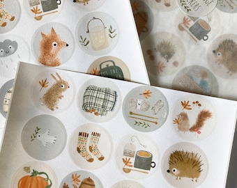 May include: A sheet of stickers featuring illustrations of woodland creatures, cozy items, and autumnal elements. The stickers are round and have a white background with a light gray border. The illustrations include a hedgehog, a fox, a rabbit, a squirrel, a teapot, a mug, a pumpkin, a blanket, and a pair of socks. The text "Cosy Hygge Stickers" is printed on one of the stickers.