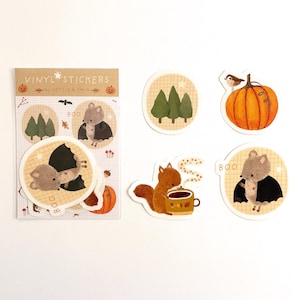 Könnte beinhalten: Eine Sammlung von Vinyl-Aufklebern mit herbstlichen und Halloween-Illustrationen. Designs umfassen ein Eichhörnchen mit einer Tasse, einen Kürbis mit einem Vogel und ein fledermausartiges Wesen. Auf dem Aufkleberbogen steht "Vinyl Stickers" und "Boo".