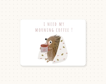 May include: A cartoon hedgehog wrapped in a blanket, holding a red mug of coffee. The text "I need my morning coffee!" is written above the hedgehog.